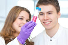 Eine Wissenschaftlerin und ein Wissenschaftler im Labor. Die Wissenschaftlerin hält ein Reagenzglas mit einer roten Flüssigkeit in der Hand.