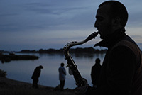 Im Vordergrund ein Saxophonist, im Hintergrund ein See in der Abenddämmerung.