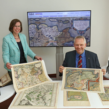 Minister Björn Thümler hält eine historische Karte in der Landesbibliothek Oldenburg hoch, im Hintergrund steht ein Bildschirm mit der digitalisierten Karte.