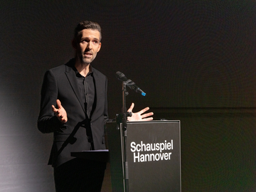 Vasco Boenisch startet in seine erste Spielzeit als Intendant des Schauspiels Hannover und hält dazu eine Rede an einem schwarzen Pult.