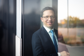 Ein Porträtfoto von Minister Falko Mohrs, in dem er lächelt und durch eine Fensterscheibe direkt in die Kamera sieht.