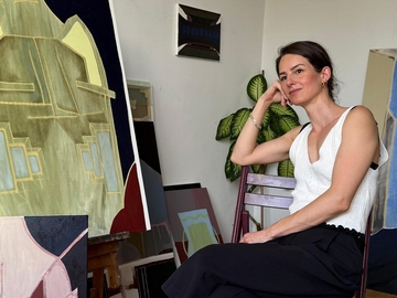 Die Künstlerin Ana Maria Szollosi ist neben einem ihrer Gemälde zu sehen. Sie stützt verträumt den Kopf auf eine Hand und schaut am Betrachter vorbei aus dem Bild heraus.