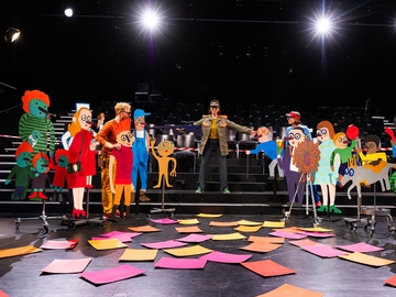 Eine Frau in Militärjacke steht auf einer Bühne, sie hat die Arme selbstbewusst ausgebreitet, als würde sie jemanden zurückweisen. Sie ist umringt von bunten Pappfiguren, die sie mit großen Kulleraugen anschauen.