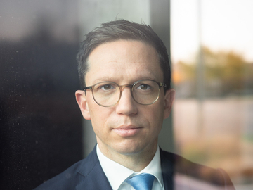 Minister Falko Mohrs schaut den Betrachter des Fotos durch eine Fensterschreibe ernst und durchdringend an. Seine Stirn ist ein Falten gelegt. Er trägt seinen klassischen dunkelblauen Anzug.