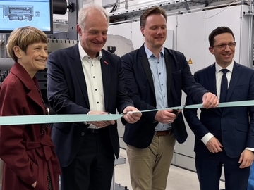 Vier Personen stehen in einer Werkhalle und schneiden gemeinsam ein mintfarbenes Band durch. Links eine Frau im roten Mantel, daneben drei Männer in Anzug oder Sakko. Hinter ihnen stehen technische Geräte und Maschinen.