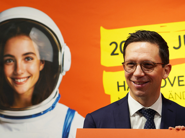 Minister Falko Mohrs steht vor einem orangefarbenen IdeenExpo‑Plakat und hält ein großes Schild mit dem Logo ‚IdeenExpo 26‘. Hinter ihm ist das Foto einer jungen Frau im Astronautenanzug auf dem Plakat zu sehen.
