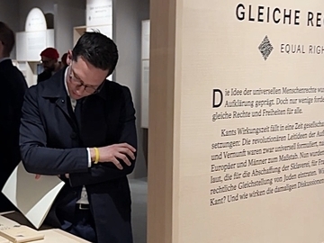Minister Falko Mohrs steht in einer Ausstellung und betrachtet nachdenklich eine Infowand mit dem Titel ‚Gleiche Rechte‘. Vor ihm liegen mehrere Bücher aus. Hinter der Wand sind weitere Besucher und Ausstellungstafeln zu sehen.