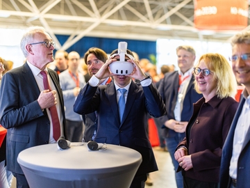 Minister Falko Mohrs probiert inmitten einer Gruppe von Besucherinnen und Besuchern eine VR‑Brille aus. Um ihn herum stehen mehrere Personen an Stehtischen und beobachten ihn lächelnd in einer hellen Veranstaltungshalle.