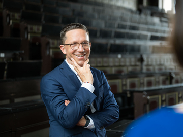 Minister Falko Mohrs steht lächelnd in einem historischen Hörsaal, den Kopf nachdenklich mit der Hand abgestützt. Er blickt einer Person im Vordergrund zu, hinter ihm steigen dunkle, hölzerne Sitzreihen steil nach oben.