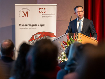 Minister Falko Mohrs hält eine Rede an einem Rednerpult mit Blumenarrangement. Neben ihm steht ein Banner mit dem Logo des Museumsgütesiegels Niedersachsen und Bremen. Im Vordergrund sitzt Publikum im abgedunkelten Saal.