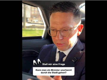 Minister Falko Mohrs sitzt auf dem Rücksitz eines Autos und blickt konzentriert auf sein Smartphone. Sonnenlicht fällt durch das Fenster. Auf dem Bild steht die Frage: ‚Kann man als Minister unerkannt durch die Stadt gehen?‘.