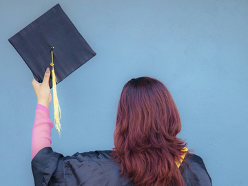 Eine Frau im schwarzen Talar steht mit dem Rücken zur Kamera vor einer blauen Wand. Sie hebt einen Graduation‑Hut mit goldener Quaste in die Höhe. Langes rötliches Haar fällt über den Talar.“