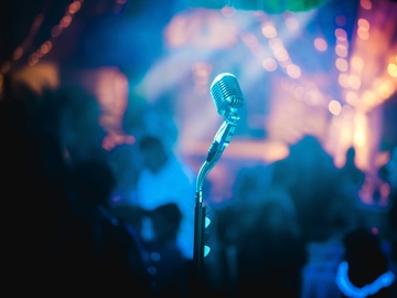 Vintage‑Mikrofon in blauem Bühnenlicht, zentral im Vordergrund. Dahinter eine unscharfe Menschenmenge in Konzert‑ oder Clubatmosphäre mit warmen, verschwommenen Lichterketten.