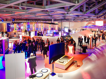 Große, moderne Ausstellungshalle mit vielen Besucherinnen und Besuchern. Zwischen futuristischen Installationen, Monitoren und beleuchteten Ständen stehen Gruppen im Gespräch. Farbenfrohes Licht in Blau, Lila und Orange prägt die offene Raumstruktur.