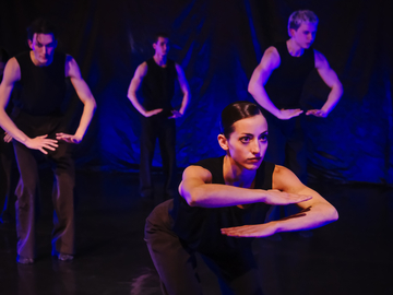 Bühnenaufnahme einer Tanzperformance: Mehrere Tänzerinnen und Tänzer in schwarzen, ärmellosen Kostümen stehen gestaffelt auf dunkler Bühne. Die vordere Person geht in die Hocke, die Arme waagerecht vor dem Körper, blaues Licht beleuchtet die Szene
