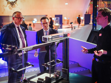 Drei Personen stehen in einer modern beleuchteten Ausstellungshalle vor einer technischen Demonstrationsanlage aus Metall und Glas. Eine Person erklärt das Exponat mit Handgesten, die beiden anderen beobachten aufmerksam.