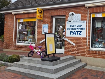 Außenansicht der Buchhandlung Patz mit roter Backsteinfassade. Über den Schaufenstern steht ‚Buch und Schreibwaren PATZ‘ mit Eulen‑Logo. Vor dem Eingang stehen Stufen, ein Aufsteller und ein Kinderfahrrad; rechts ein gelber Briefkasten.