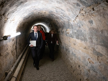 Minister Falko Mohrs geht gemeinsam mit mehreren Personen durch einen schmalen, gemauerten Gewölbetunnel. Entlang der linken Wand verlaufen Rohre und eine Leuchte. Der Boden ist gekiest, die Tunnelwände sind rau und hellgrau.