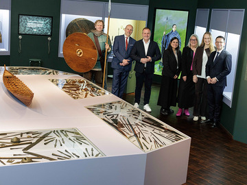 Heller Museumsraum mit großer, weißer Tischvitrine, in der zahlreiche eiserne Waffen und Werkzeuge liegen. Darauf steht ein Holzmodell eines Bootes. Dahinter steht eine Gruppe von Personen vor Wandvitrinen und lebensgroßen Figuren mit Schild und Speer