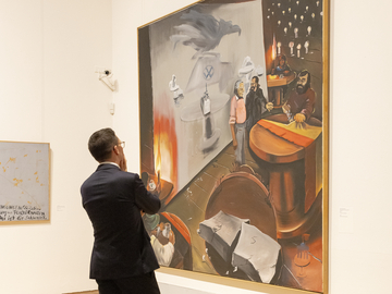 Minister Mohrs steht in einem hellen Museumsraum vor einem großformatigen Gemälde. Das Bild zeigt eine rätselhafte Szene mit mehreren Figuren an einem Tisch, Kerzenlicht, dunklen Farben und surreal wirkenden Motiven.