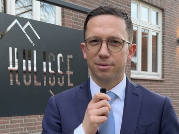 Minister Falko Mohrs steht im Freien vor einem Backsteingebäude und dem Schild ‚KULISSE‘. Er trägt Anzug und Krawatte und hält ein kleines Mikrofon in der Hand. Hinter ihm sind Fenster und eine niedrige Steinmauer zu sehen.