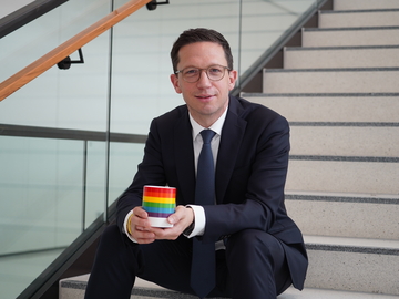 Minister Mohrs sitzt auf der Treppe im Landtag und hält eine Tasse mit der Regenbogenflagge in den Händen