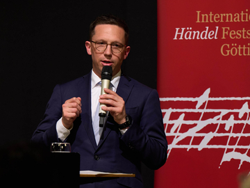 Minister Mohrs hält eine Rede auf einer Bühne, hinter ihm ist das rote Banner der Internationalen Händel Festspiele in Göttingen zu sehen.