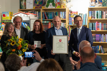 Die Familie Patz mit der Urkunde zum Buchhandelspreis 2026 und Minister Mohrs. Sie stehen vor einem bunten Bücherregal in ihrem Laden. Einige Zuschauende fotografieren.