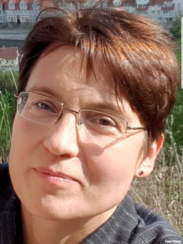 Nahaufnahme einer Person mit kurzer Frisur und schmaler Brille im Freien. Im Hintergrund sind unscharf Häuser, Vegetation und ein Hang zu erkennen.