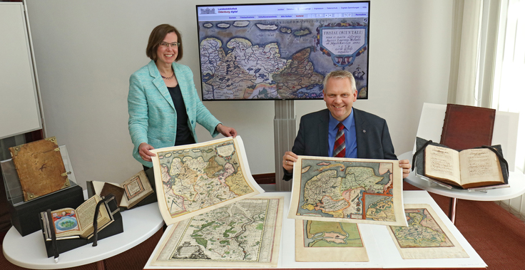 Minister Björn Thümler hält eine historische Karte in der Landesbibliothek Oldenburg hoch, im Hintergrund steht ein Bildschirm mit der digitalisierten Karte.