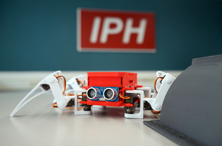 Eine kleine rote Roboterspinne steht vor dem IPH-Logo. Das Logo ist verschwommen im Hintergrund.