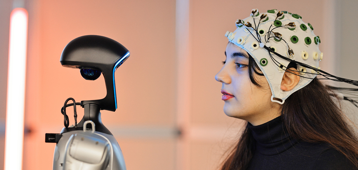 Eine junge Frau mit einer Elektrodenhaube auf dem Kopf betrachtet einen Roboter.
