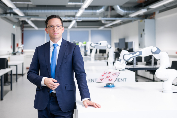 Minister Falko Mohrs steht in einem weißen Labor voller Roboterarme und schaut direkt in die Kamera.