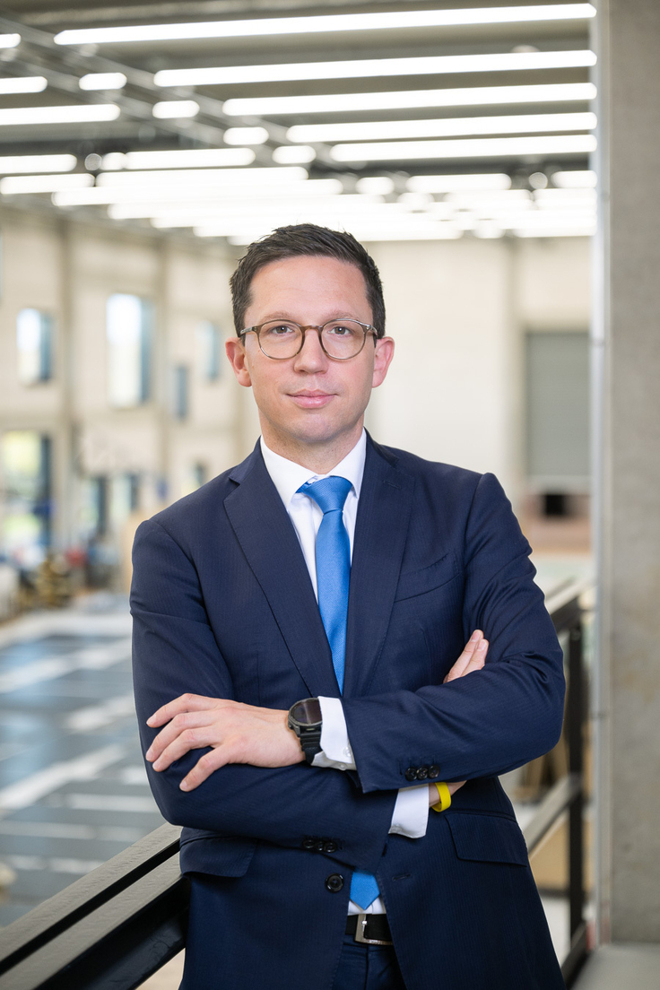 Minister Falko Mohrs steht mit verschränkten Armen in einer Werkstatt. Er schaut mit erstem Gesichtsausdruck direkt in die Kamera.