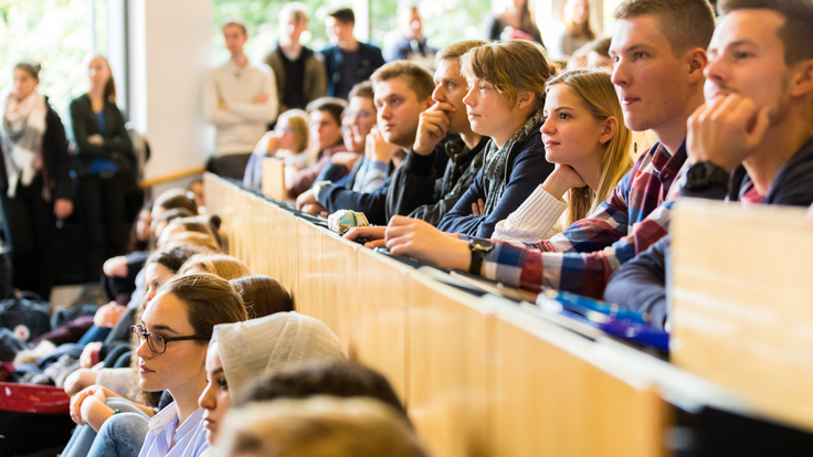 Auf dem Foto sitzen Studierende in einem vollen Hörsaal und hören aufmerksam zu. Wer zu in ihnen spricht, ist nicht zu erkennen.