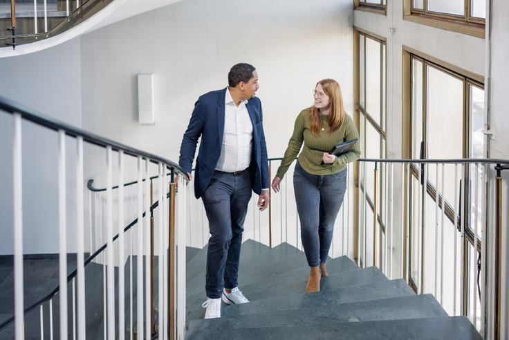 Eine junge Frau und ein junger Mann gehen die Treppe im MWK hoch und unterhalten sich lachend.