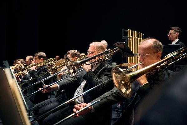 Das Foto zeigt sechs Männer in Anzügen, die professionell Posaune spielen. Im Hintergrund steht ein Mann am Percussions-Ensemble.