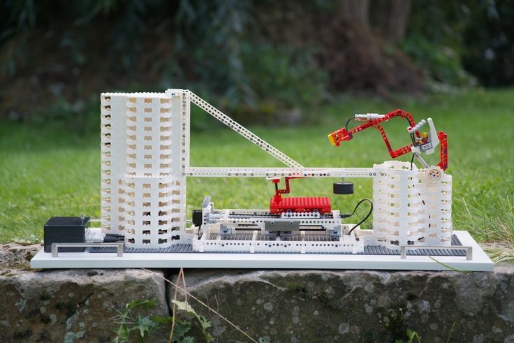 Nachbau eines Rasterkraftmikroskops aus Klemmbausteinen: zwei weiße zylindrische Türme, verbunden durch eine filigrane Brücke. Rechts ein beweglicher roter Roboterarm. Das Modell steht draußen auf einer Steinfläche vor unscharfem Grün.
