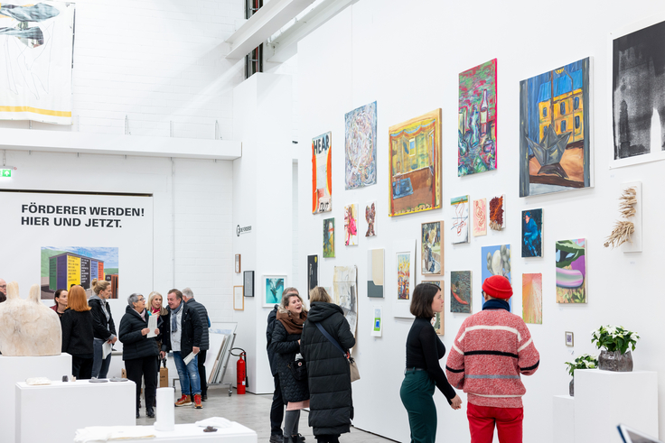 Innenansicht eines hellen Museumsraums mit hohen weißen Wänden. Viele kleine und große, farbige Gemälde hängen dicht nebeneinander. Mehrere Besucher stehen und gehen, betrachten die Kunst.