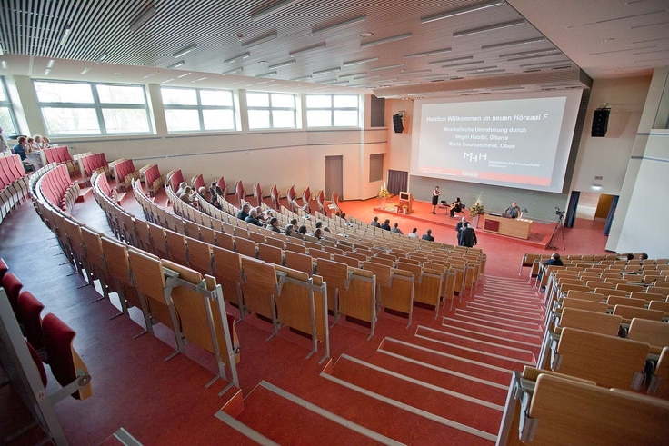 Ein leerer Hörsaal mit roten Stühlen und einer Projektionsfläche im Hintergrund.