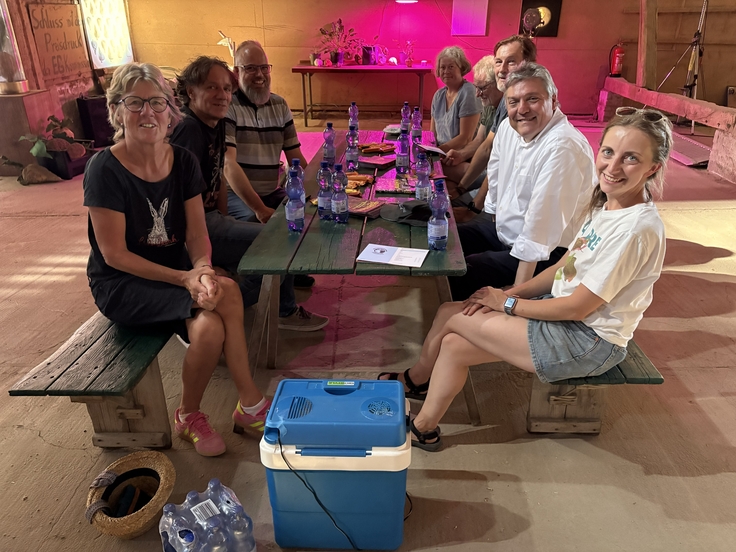 Acht Personen sitzen an einem langen Holztisch in einem Veranstaltungsraum mit farbiger Beleuchtung. Auf dem Tisch stehen Wasserflaschen und Unterlagen. Im Vordergrund sind eine Kühlbox und Getränkekisten zu sehen, im Hintergrund eine kleine Bühne.