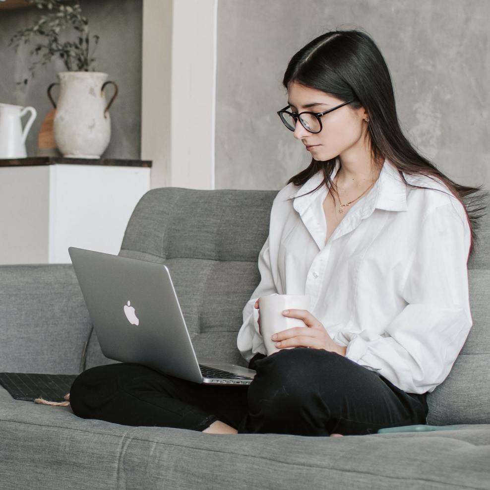 Schmuckbild: Eine junge Frau mit Brille sitzt im Schneidersitz auf einem Sofa, sie hält einen Becher in den Händen und schaut auf ein Laptop auf ihrem Schoß.
