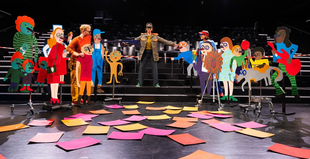 Eine Frau steht in selbstbewusster Pose mit ausgebreiteten Armen auf einer Theaterbühne. Sie trägt eine olivgrüne Militärjacke mit passender Mütze. Dabei wird sie umringt von bunten Pappfiguren, die sie mit erstauntem Ausdruck ansehen.