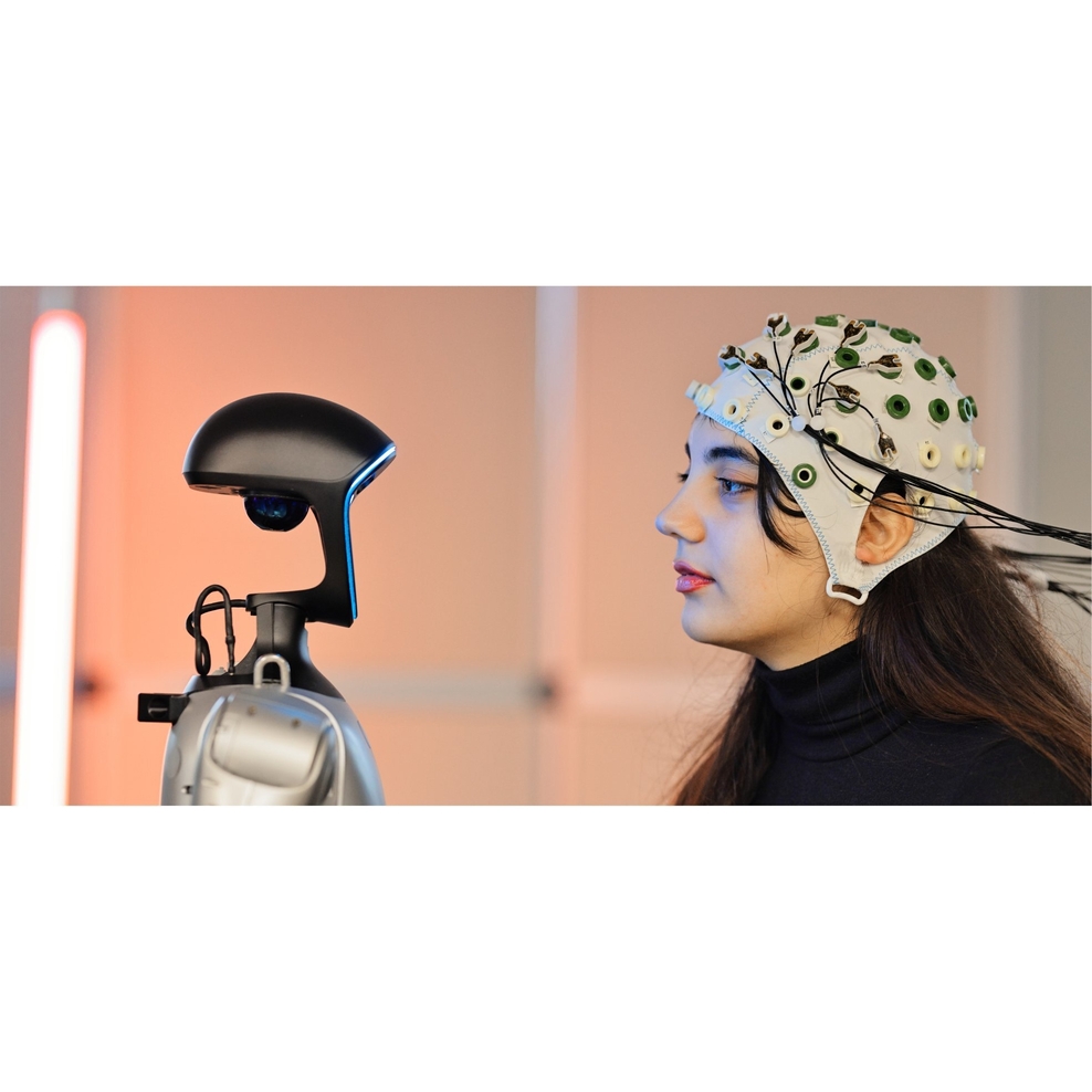 Eine junge Frau trägt eine Elektrodenhaube und schaut einen humanoiden Roboter an. Beide sind im Profil zu sehen.