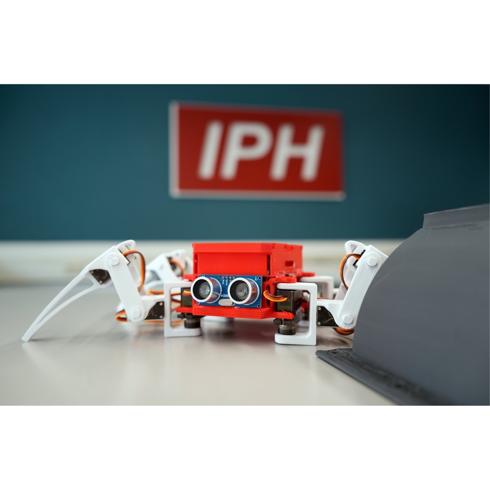 Eine rote Roboterspinne schaut aus dem Bild heraus. Hinter ihr ist verschwommen das rote IPH-Logo zu erkennen.