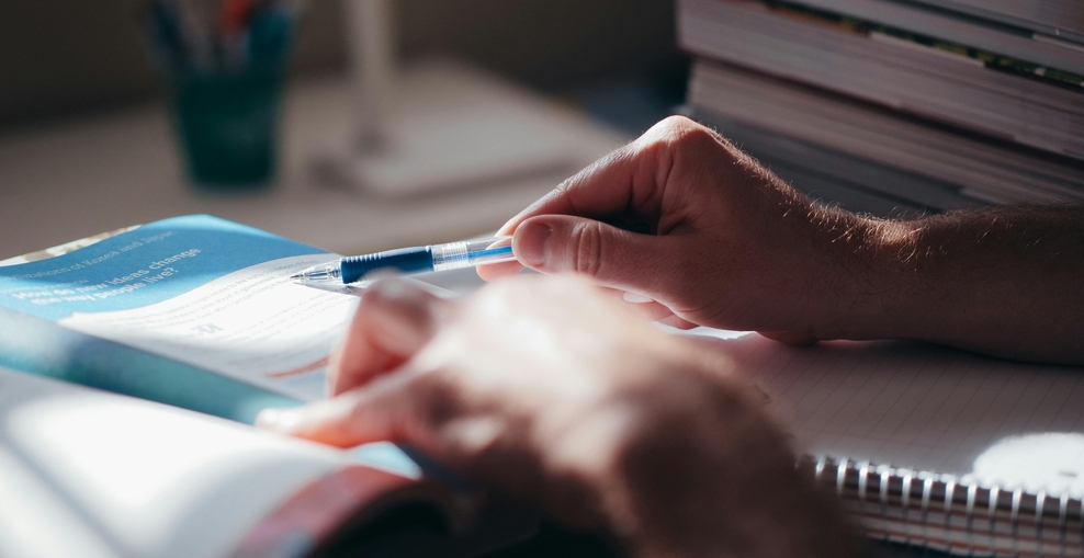 Zu sehen ist eine menschliche Hand, die mit einem Kugelschreiber auf eine Zeile in einem Buch deutet. Das Bild ist eher dunkel, im Hintergrund ist verschwommen ein Stapel Bücher zu erkennen.