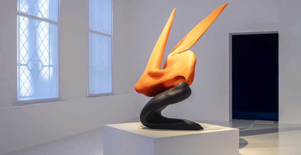 Das Bild zeigt die orange-schwarze Statue Self-Portrait von Teresa Solar Abboud in einem weißen Raum. Die Form der Statue erinnert an einen Vogel, der im Landeanflug die Flüge ausstreckt.