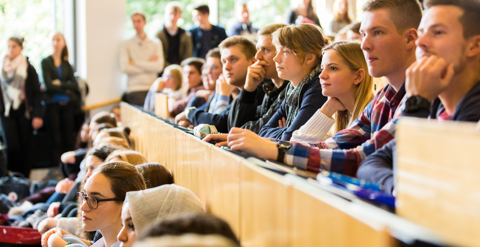 Auf dem Foto sitzen Studierende in einem vollen Hörsaal und hören aufmerksam zu. Wer zu in ihnen spricht, ist nicht zu erkennen.