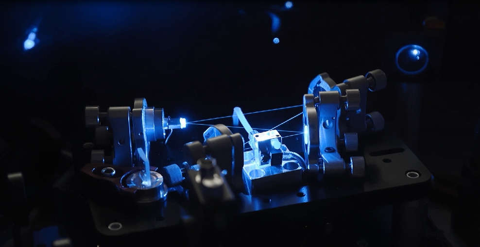 Eine Maschine schießt blaue Laserstrahlen durch einen nahezu völlig dunklen Raum. Der Hintergrund ist komplett schwarz.