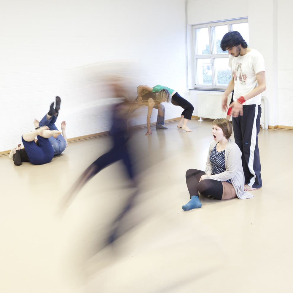 Mehrere Personen bewegen sich performativ, teils verwischt in Bewegung. Einige liegen oder sitzen am Boden, andere stehen an den Wänden. Großes Fenster, weiße Wände und viel freie Fläche.